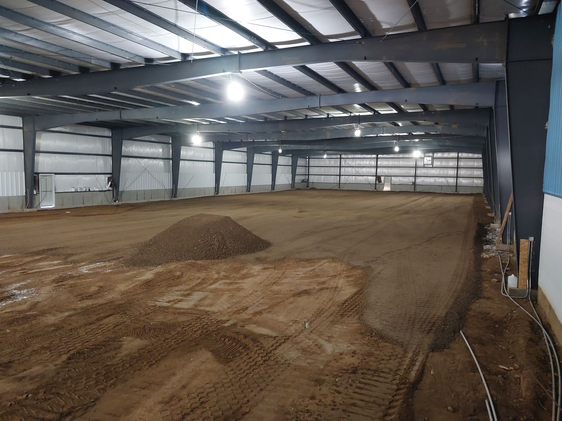 Indoor dirt arena under construction with metal frame and lighting.