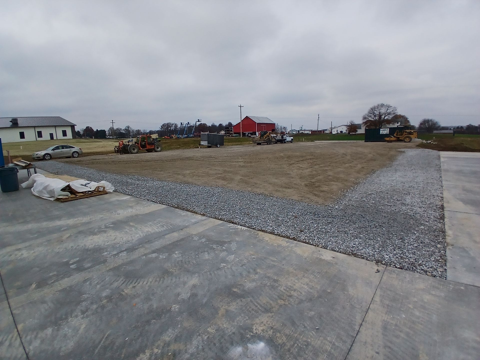 Construction site with gravel and dirt, concrete slab in foreground, red barn in the distance.