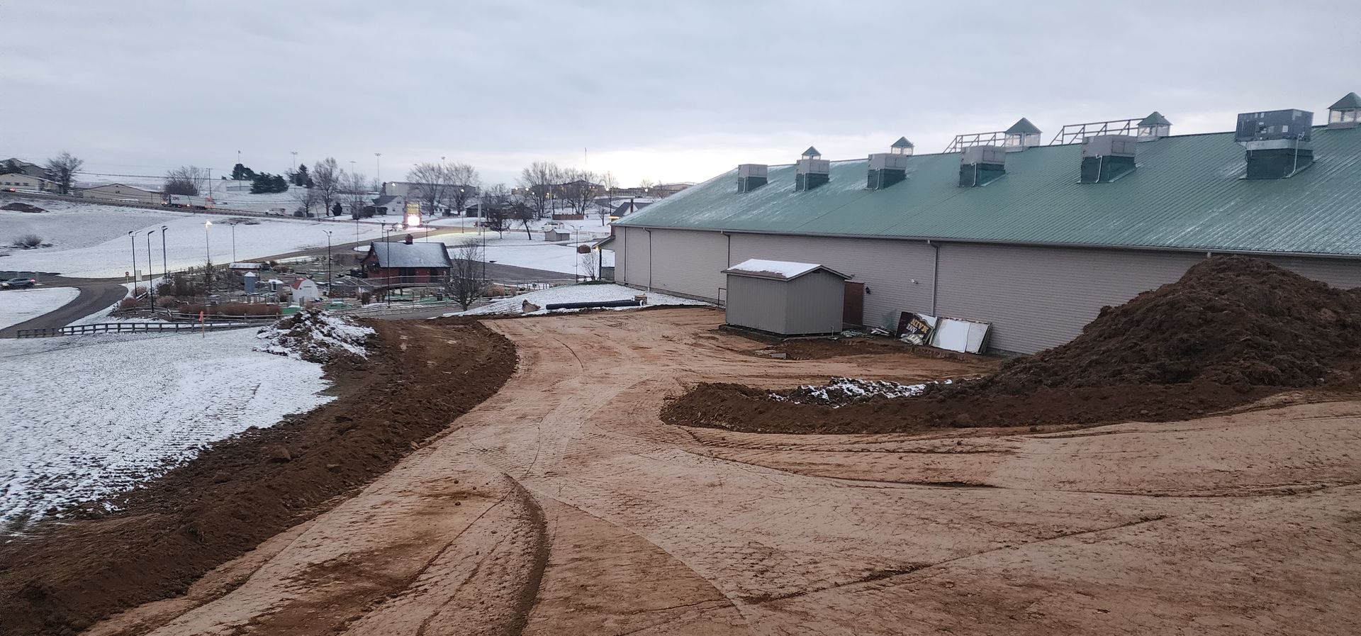 A snow-covered rural scene with a large building, a muddy foreground, and a cloudy sky.
