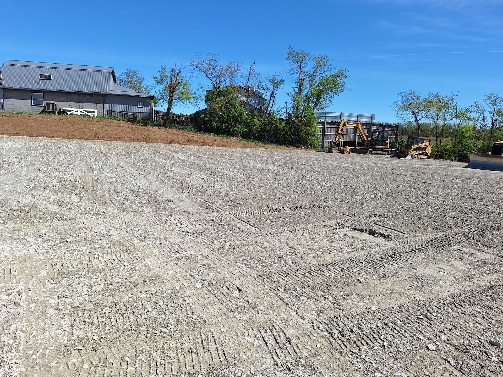 Gravel lot with tire tracks, construction vehicles in the background, a building and blue sky.