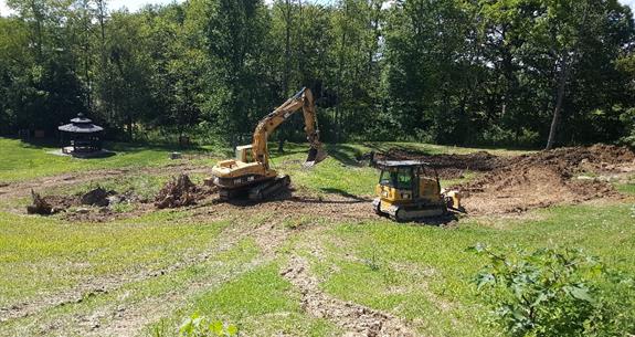 Two yellow construction excavators on a grassy field, digging near a treeline and a small structure.