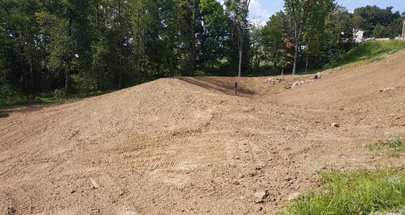 Dirt mound with graded surface, trees in the background, and grass in the foreground.