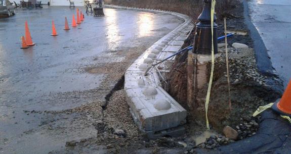 Construction site with damaged concrete curb; orange cones line road.