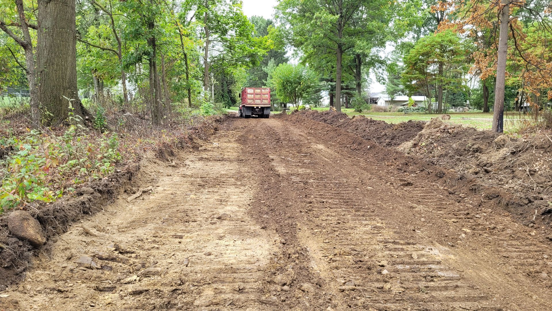 Dirt road being graded with a truck, trees in the background.