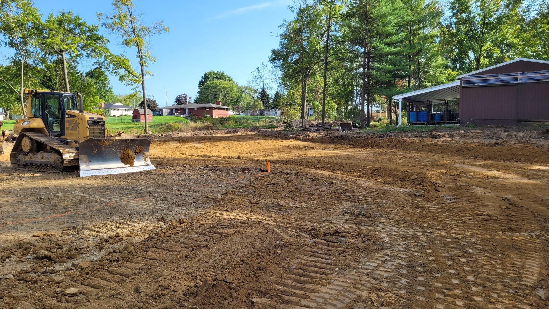 Bulldozer on a dirt lot, leveling the ground for construction. Trees and buildings in the background under a blue sky.