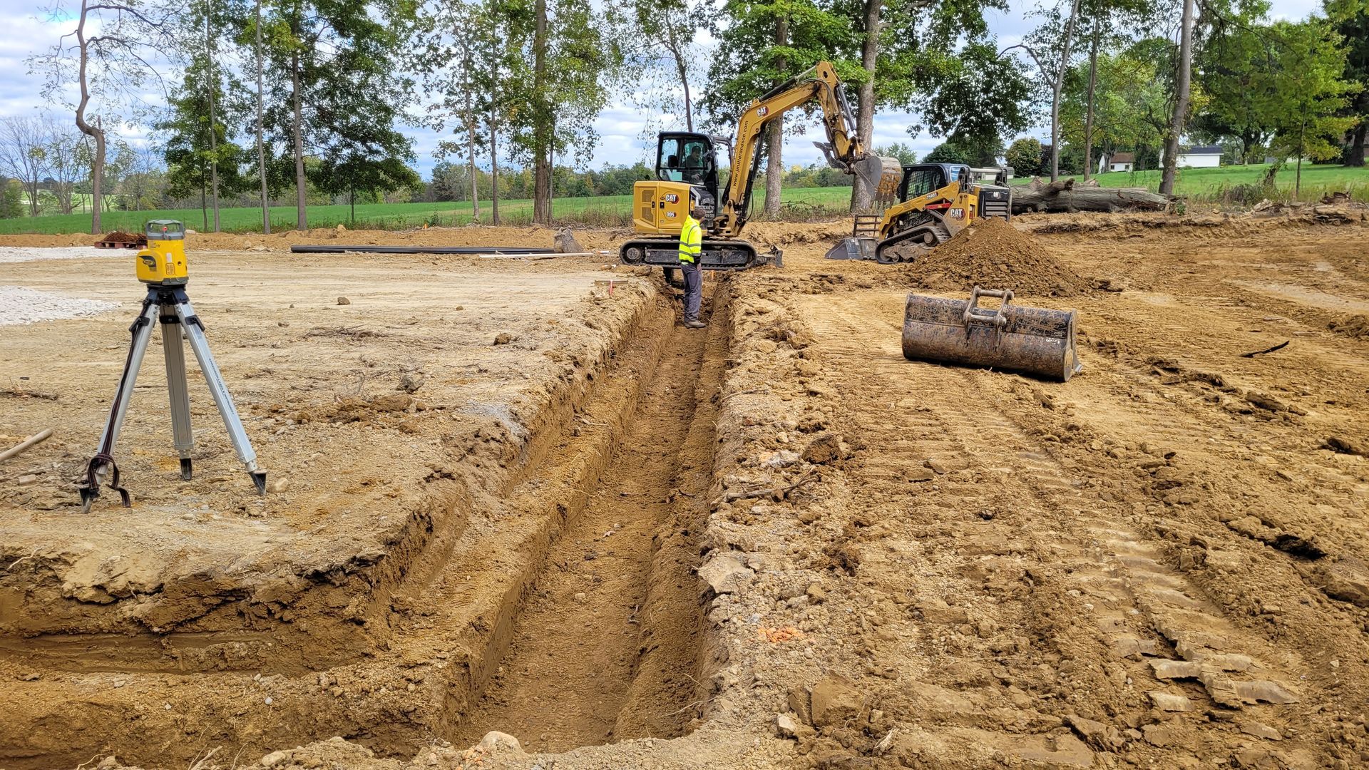 Construction site with backhoe digging trench; laser level on tripod.