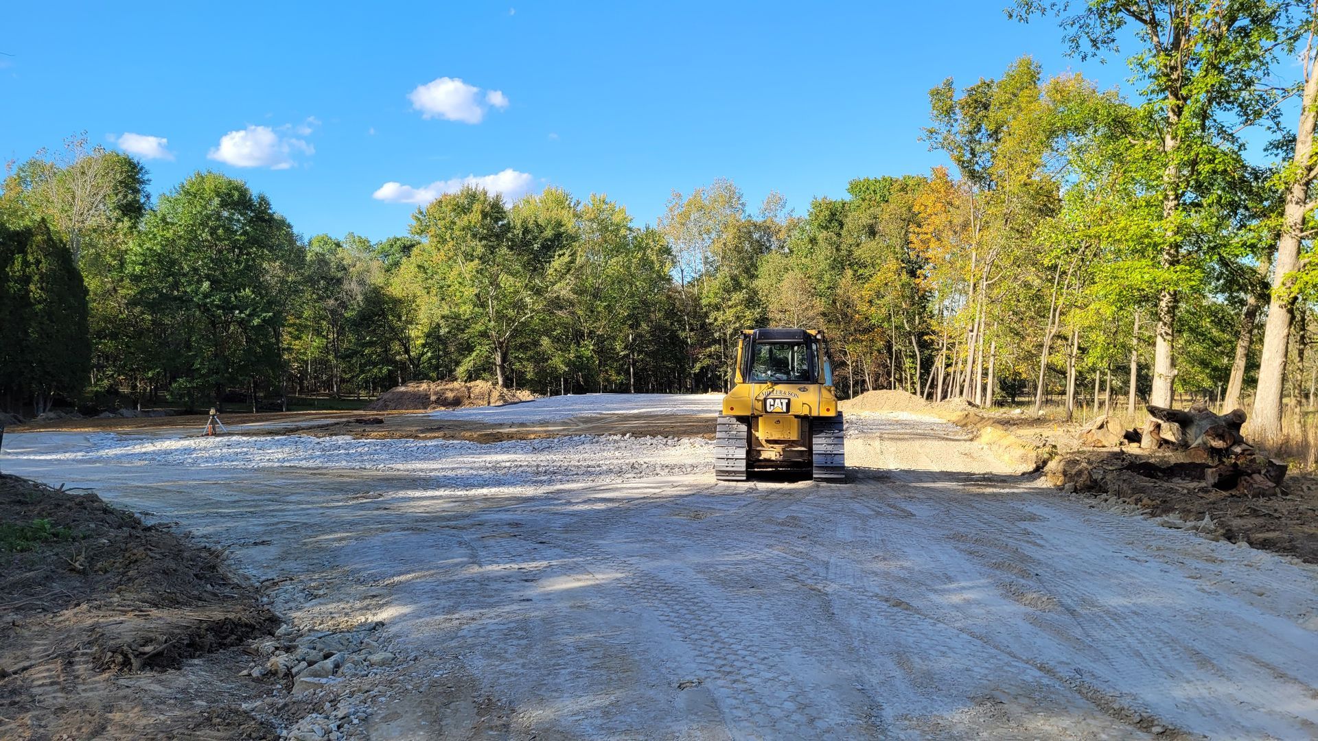 Bulldozer on a gravel path, clearing a wooded area under a blue sky.