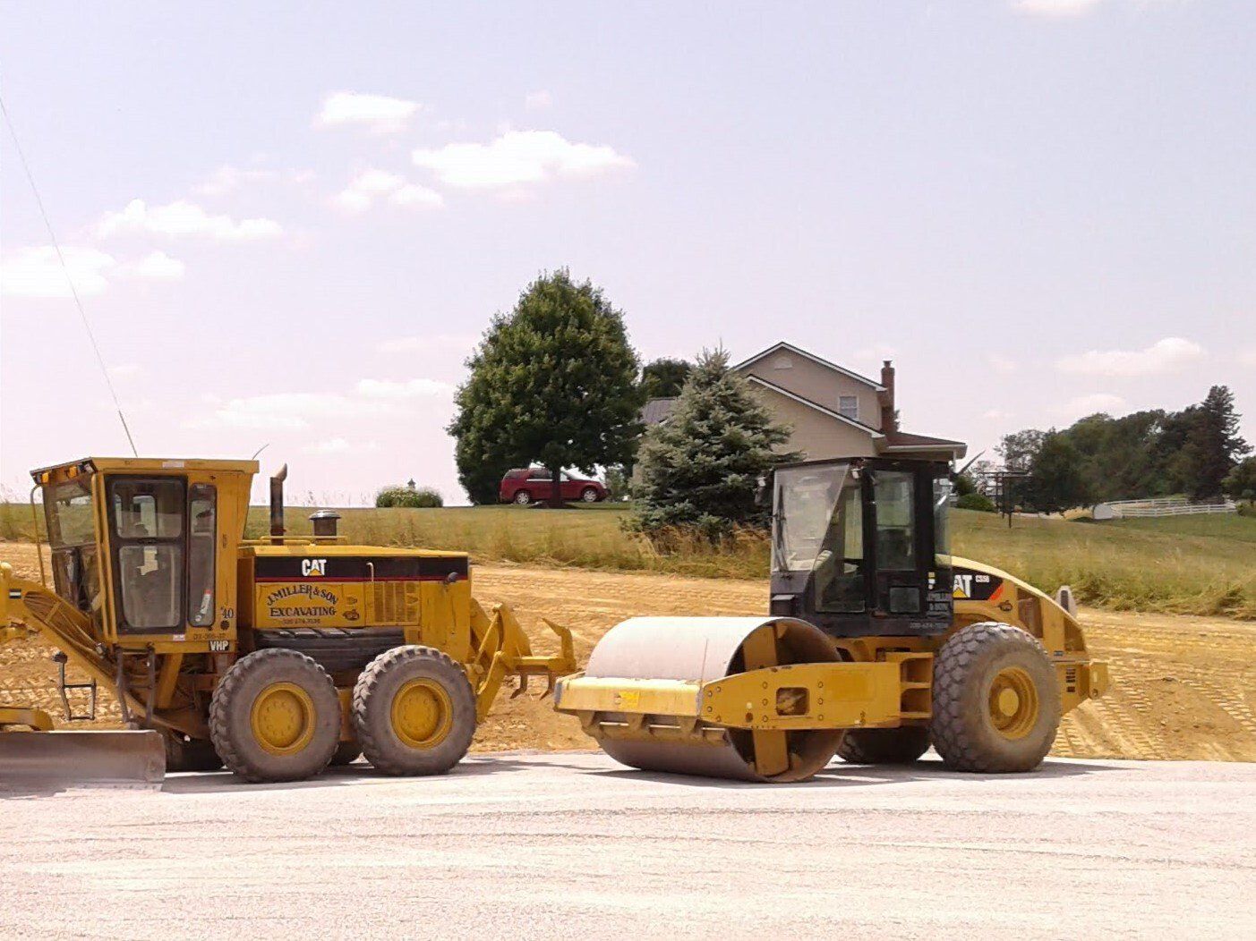 Yellow construction grader and roller on gravel, near a house and trees, under a sunny sky.