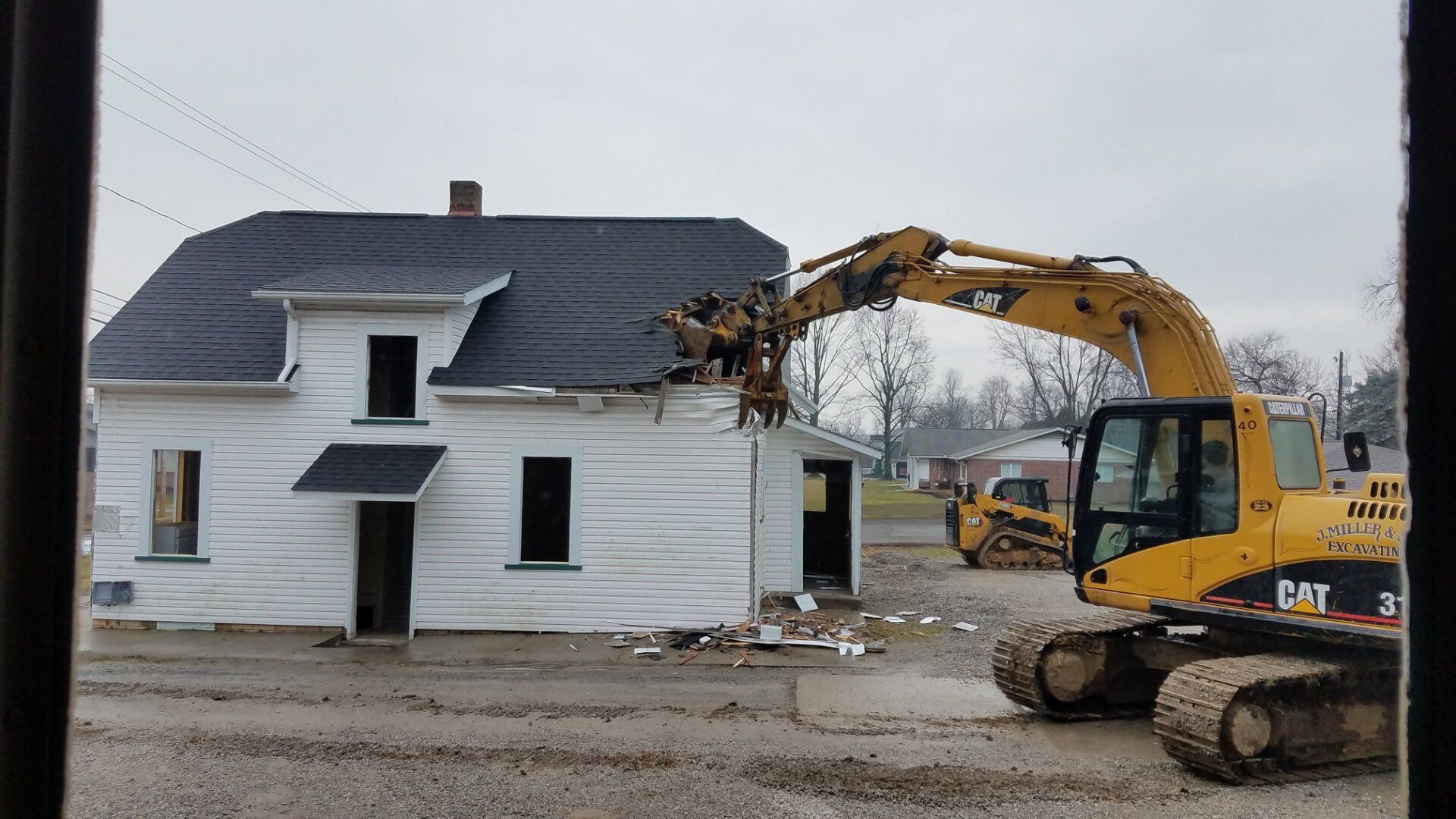 A yellow excavator demolishes a white house on a muddy lot; other construction equipment in the background.