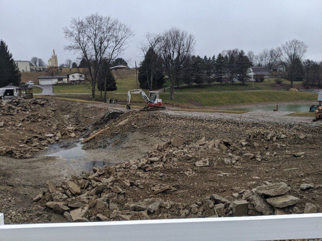 Construction site with excavator; debris, bare trees, and buildings in the background.