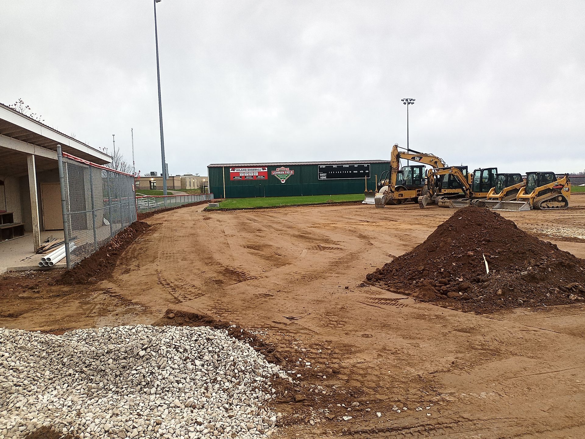 Construction site: dirt field, heavy machinery, building in background, overcast day.