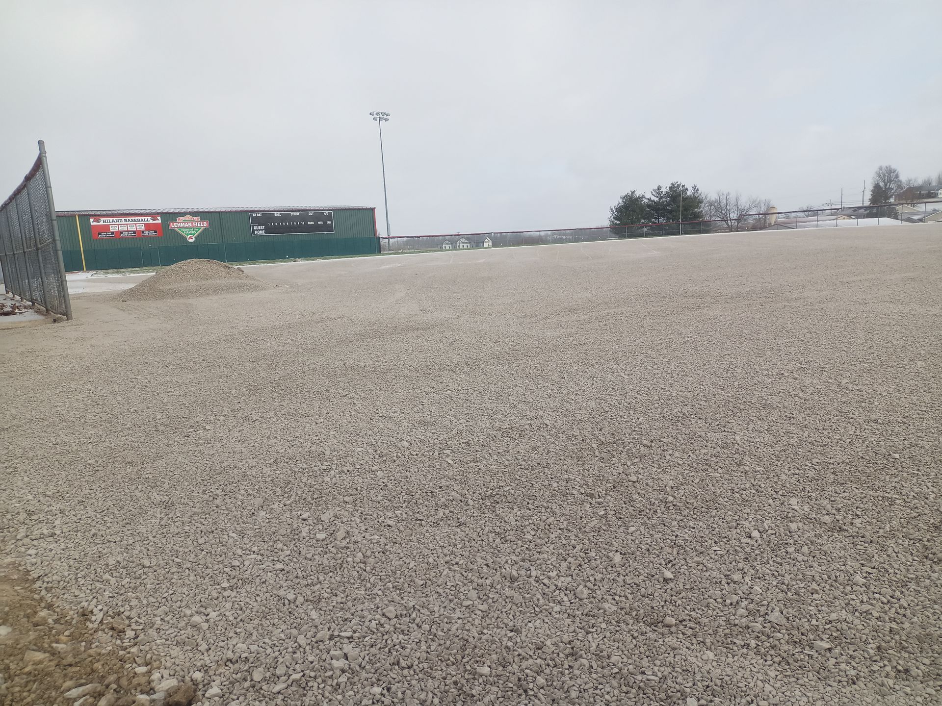 Large pile of gravel in an outdoor setting, with buildings and a tower in the background. Overcast, gray sky.