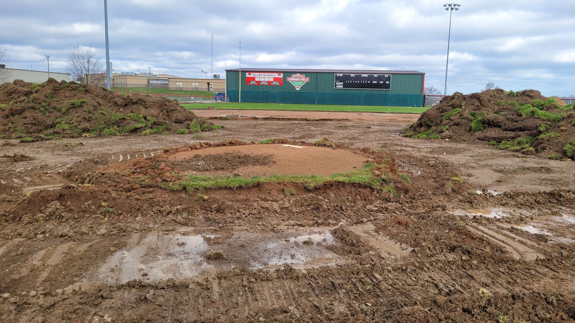 Muddy baseball field with piles of dirt, a scoreboard, and a green building in the background under a cloudy sky.