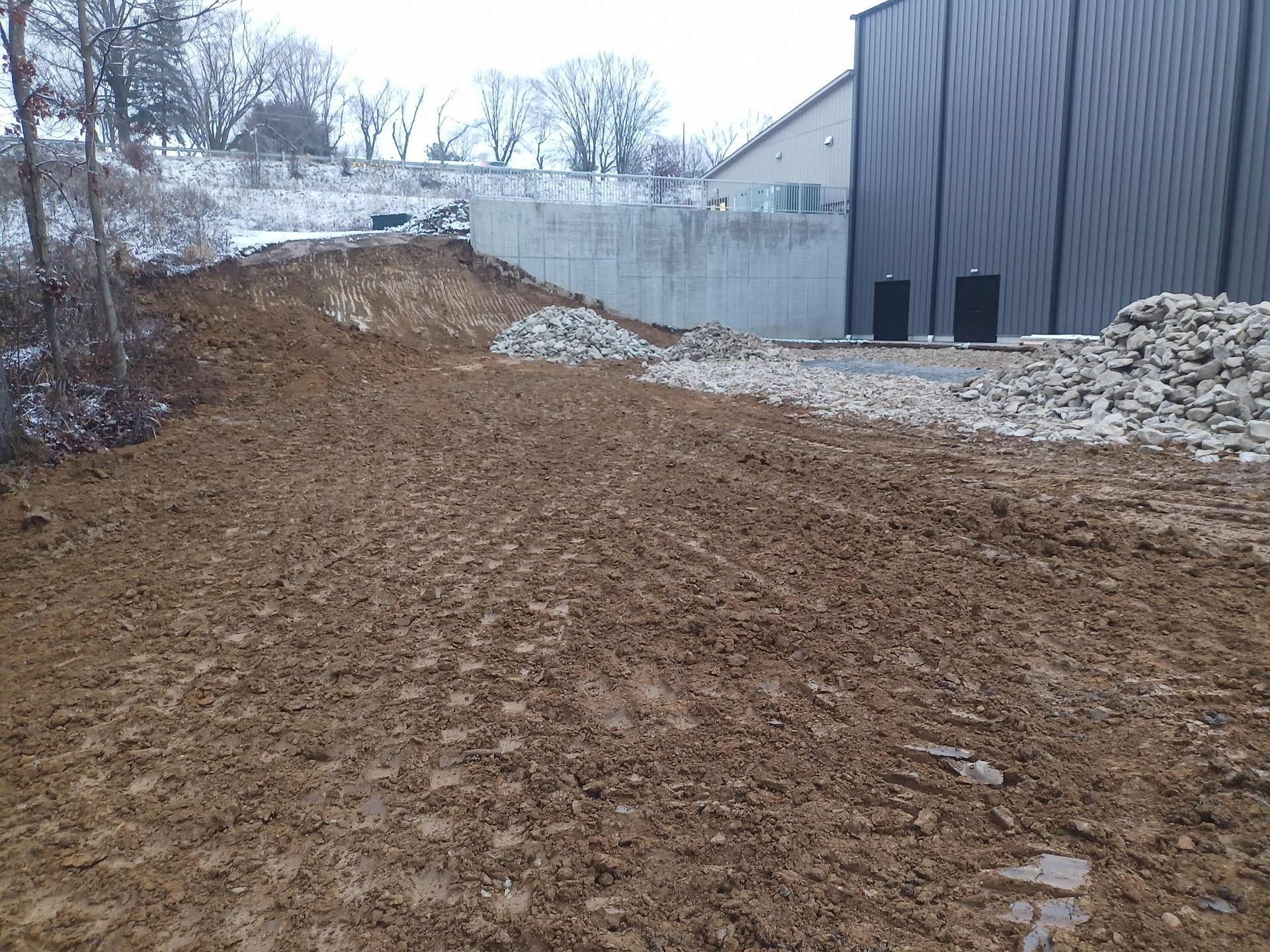 Muddy ground in front of a building and concrete wall, snow-covered trees in the background.