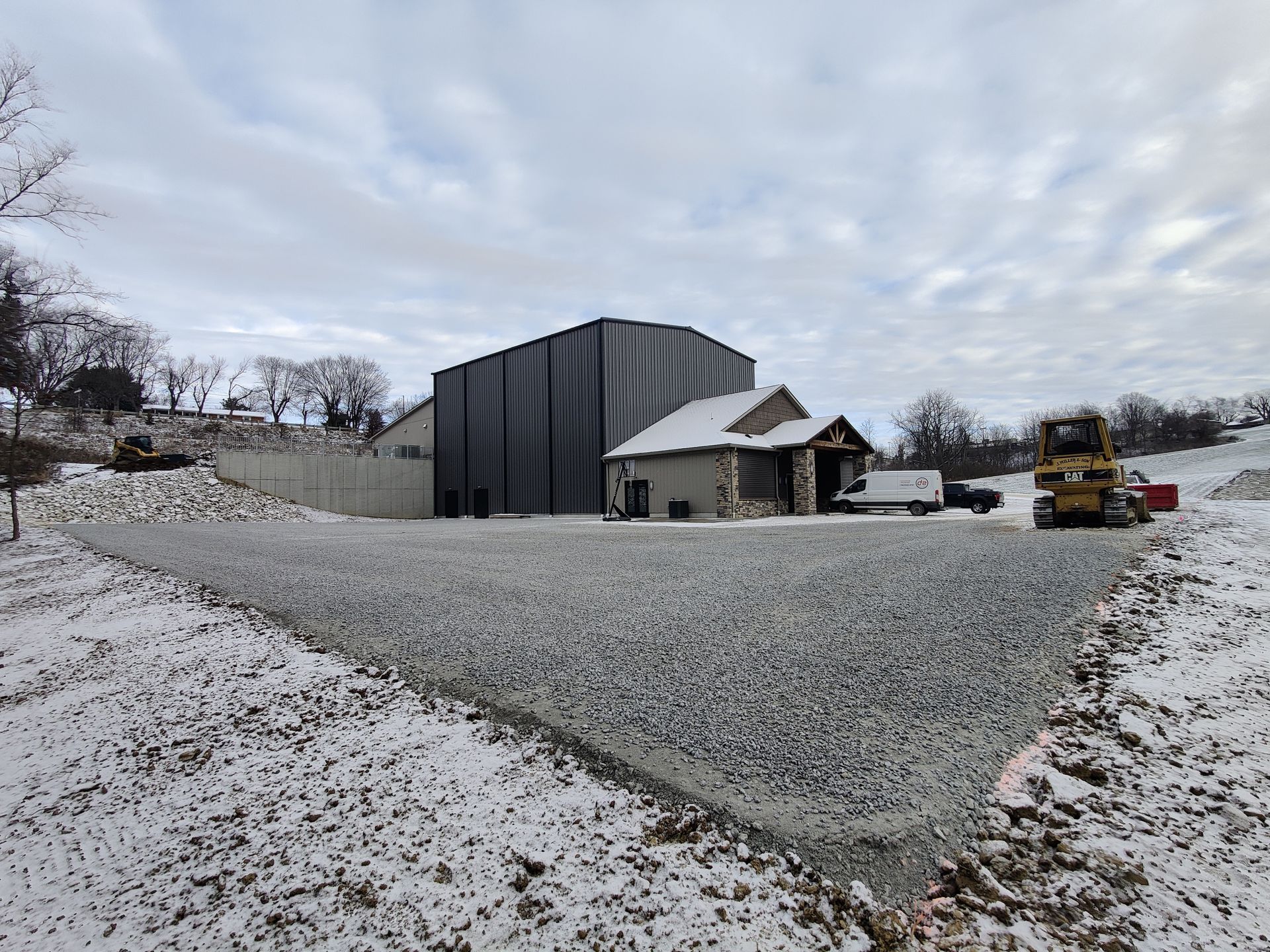 A snowy construction site with a large metal building and stone structure under a cloudy sky.
