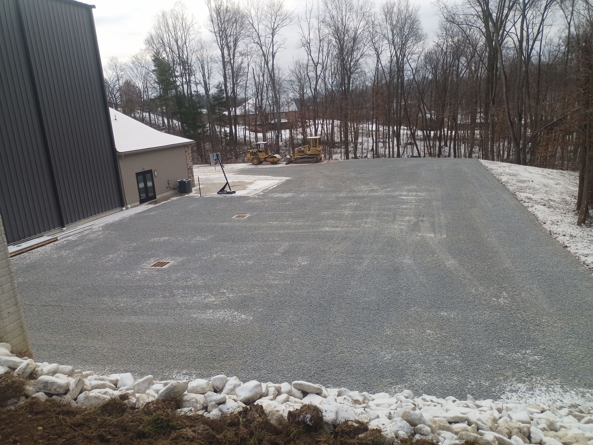Gravel-covered lot next to a building, with a few trees and snow patches. Construction equipment visible in background.