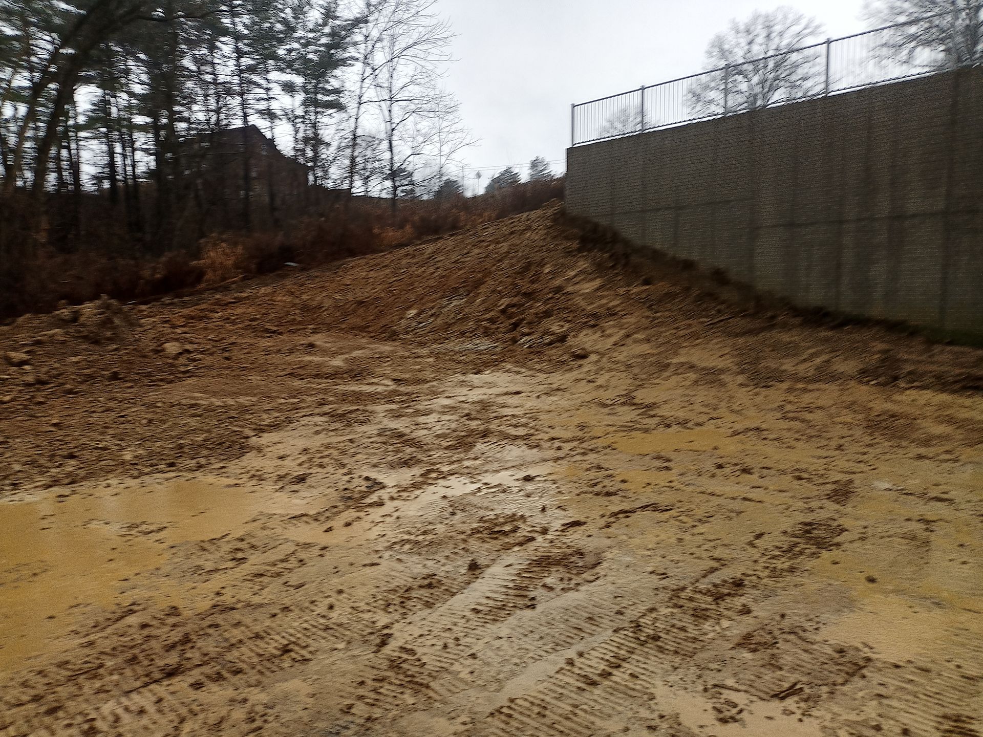 Muddy ground with a dirt pile and a retaining wall. A chain-link fence sits atop the wall.