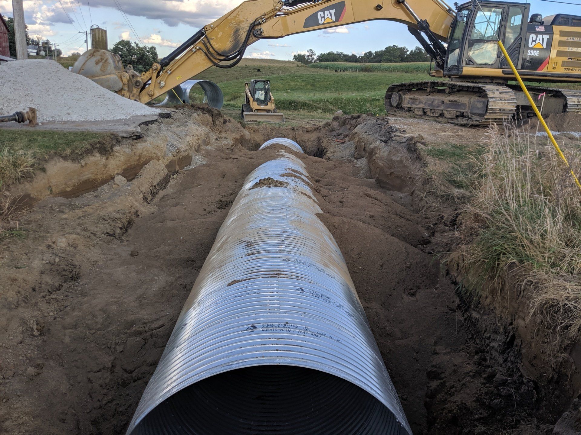 Construction site: a large corrugated metal pipe in a trench; excavator and small loader in background.