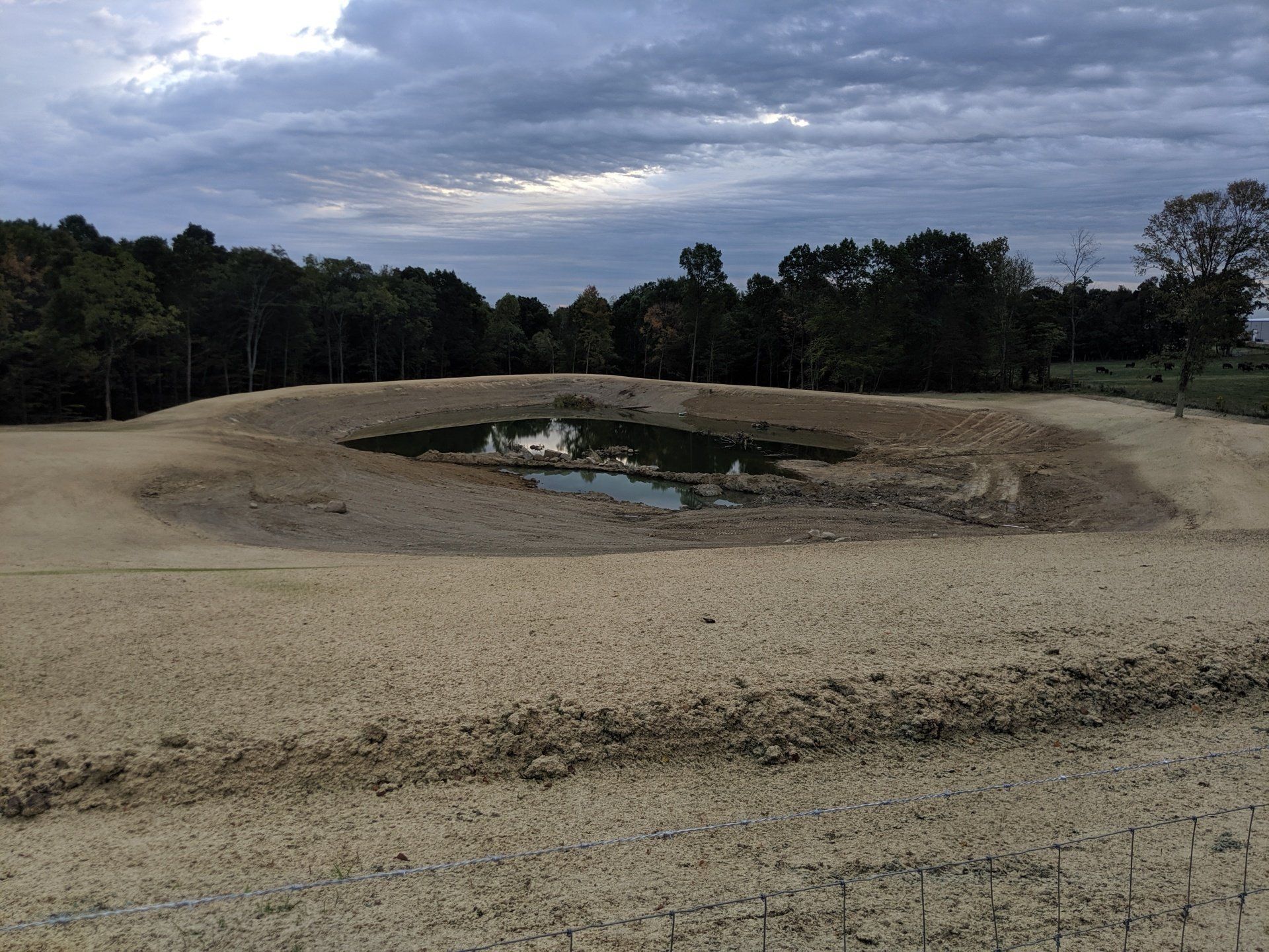 Dry pond with a small amount of water surrounded by tan, barren land and trees under a cloudy sky.