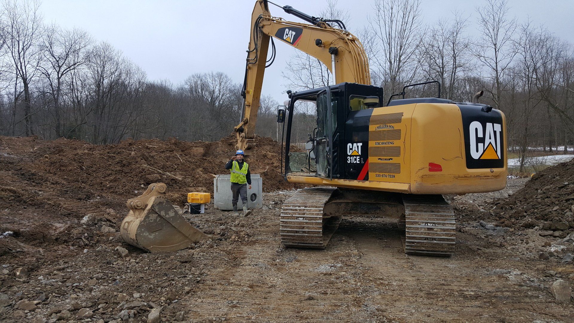 Yellow CAT excavator digging in dirt with a worker in a safety vest nearby. Cloudy sky, trees in the background.