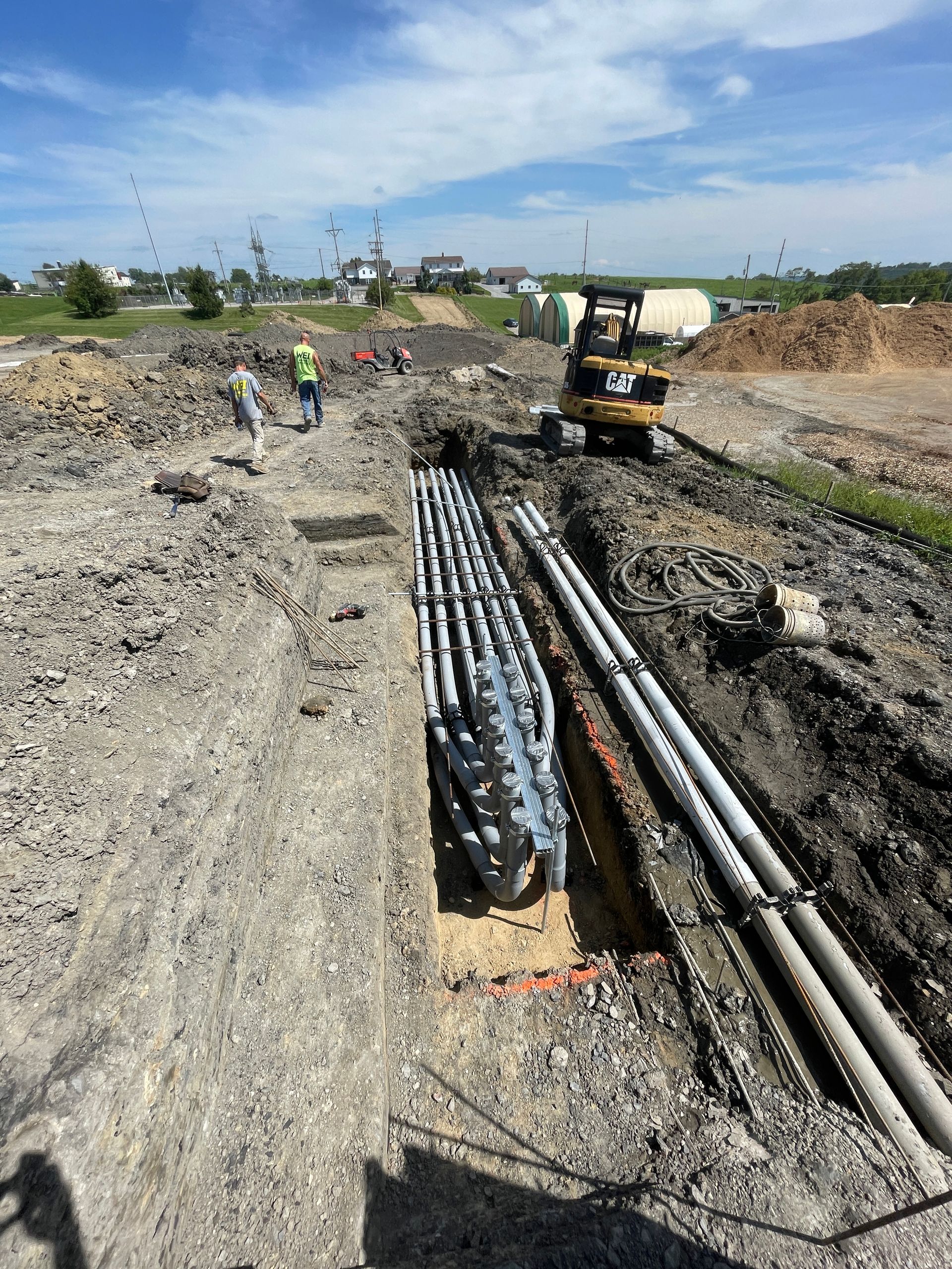 Construction site with workers, a backhoe, and conduit in a trench. Blue sky with a few birds.