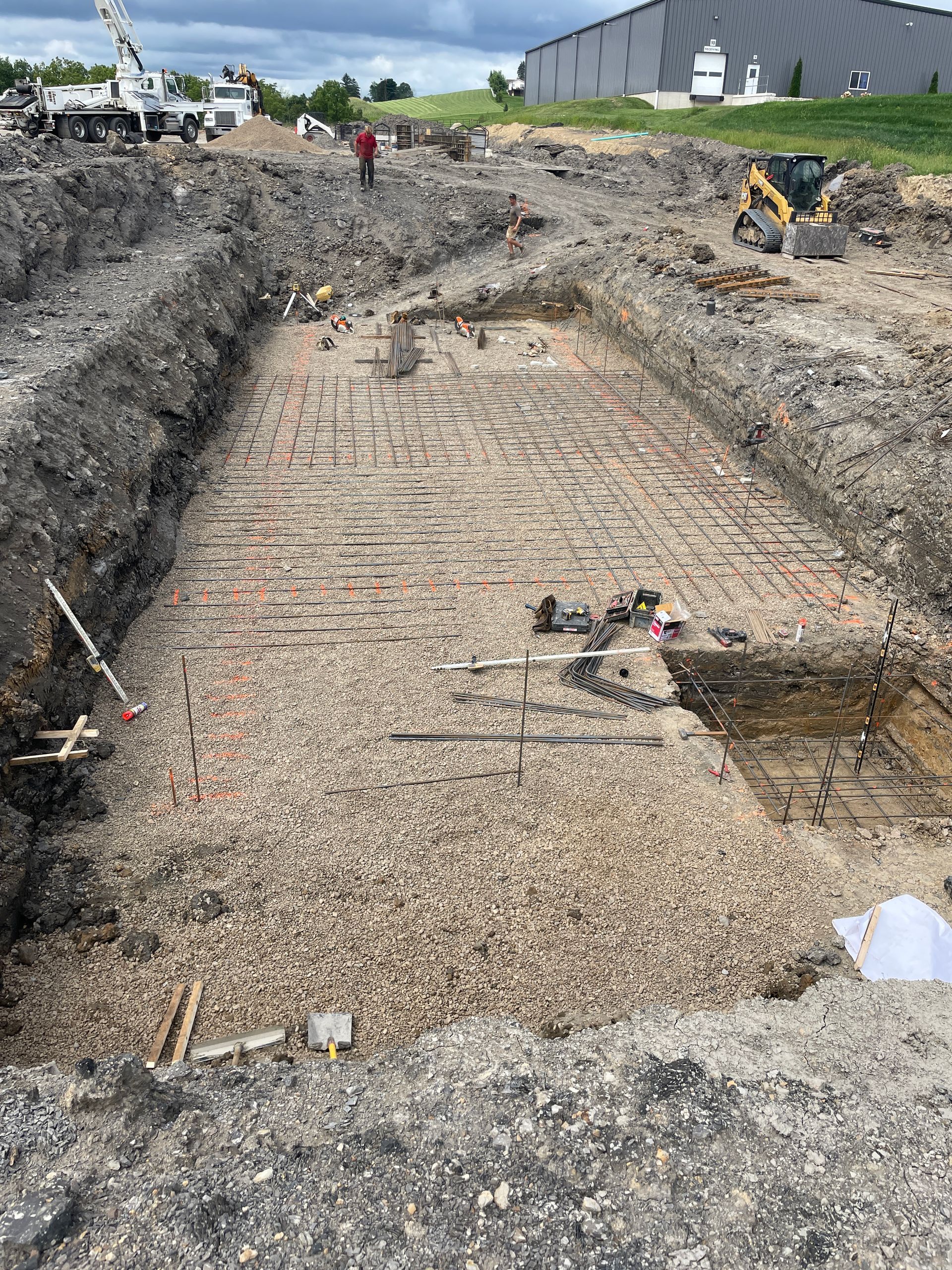 Construction site with excavated foundation. Rebar laid for concrete. Gravel base, a worker, and equipment.