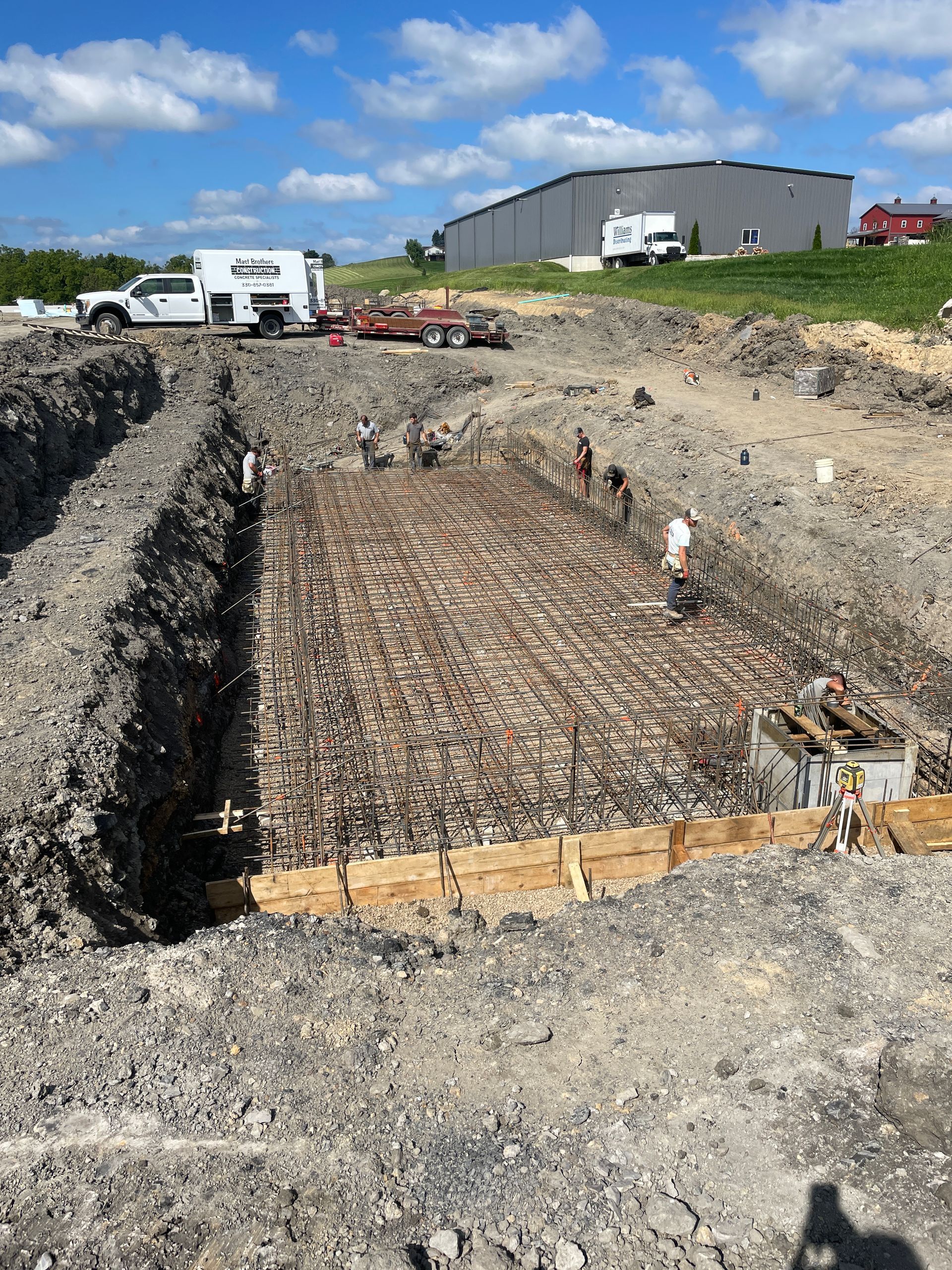 Construction site, workers around rebar framework in excavated foundation. Truck and building visible in background.