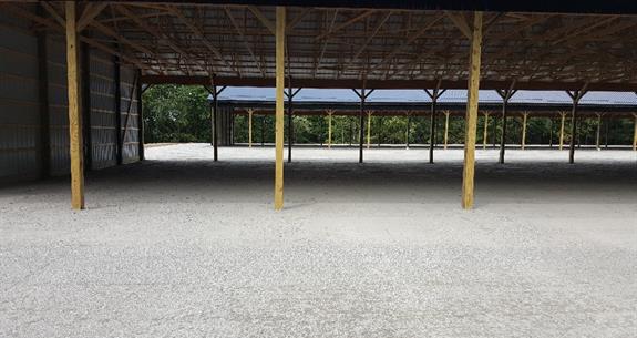 Open-air structure with gravel floor and wooden support beams. Brown roof, surrounded by green trees.