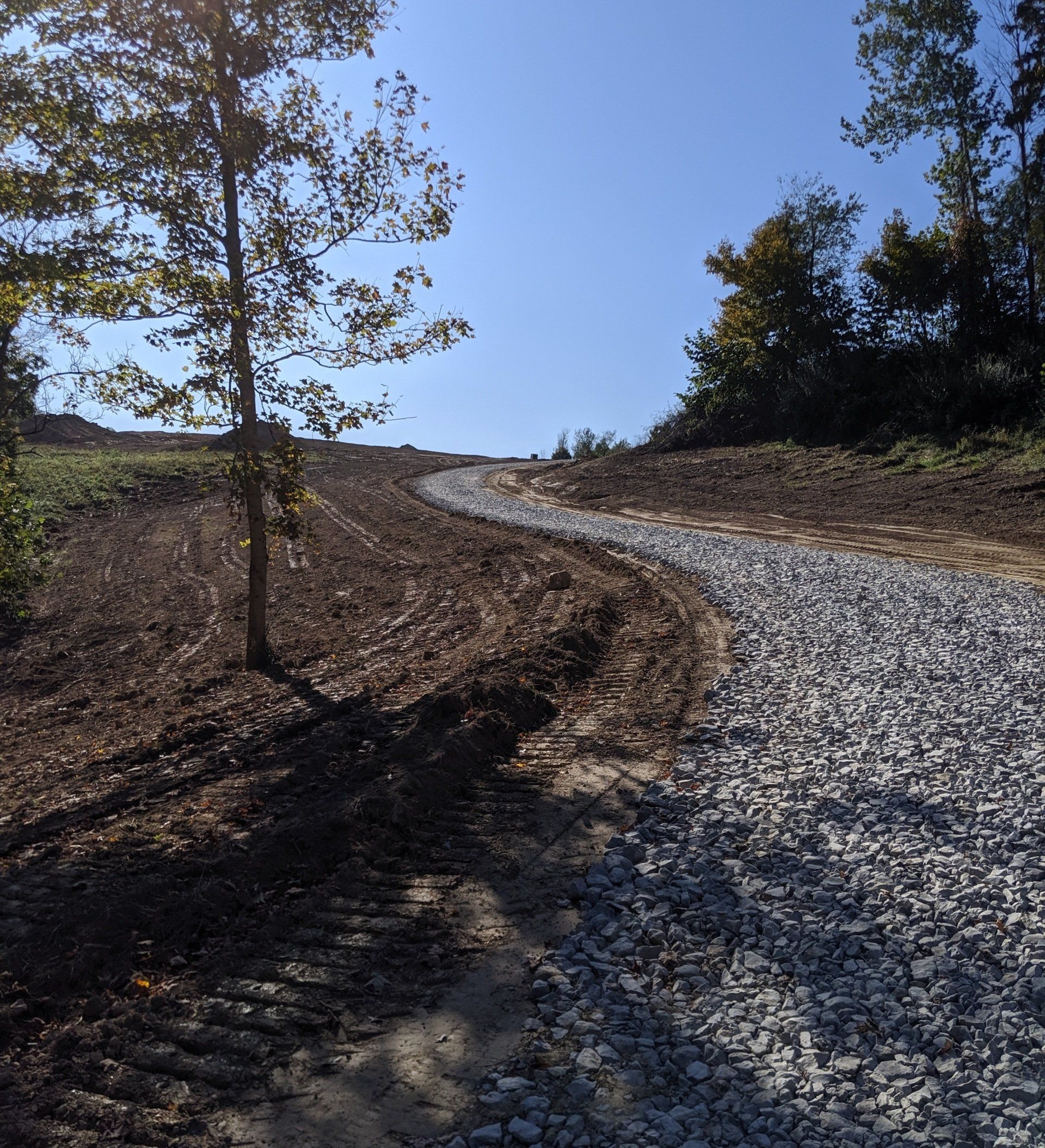 Gravel path ascending a hill. Brown soil and a few trees on either side, blue sky overhead.
