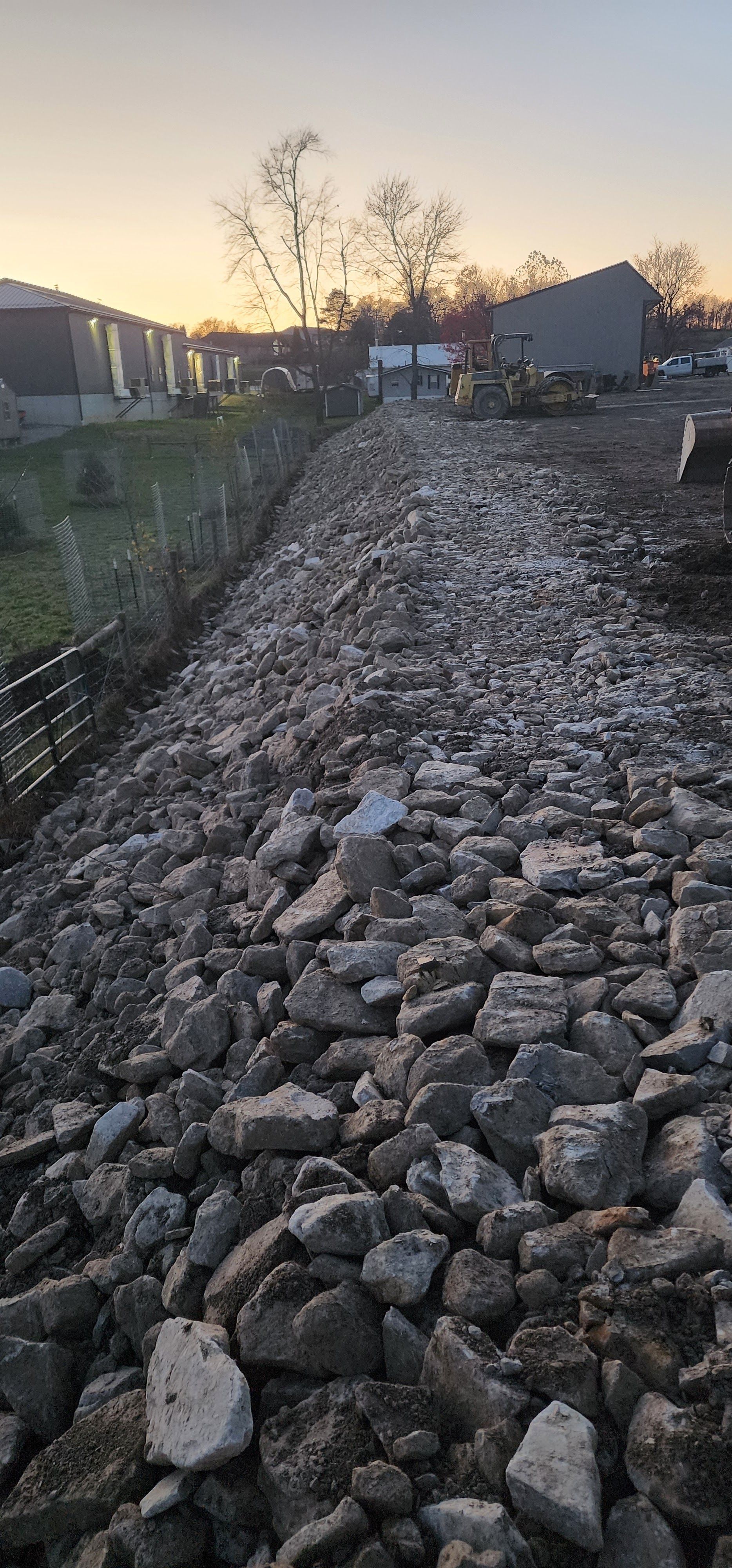 A long, rough gravel path with buildings and a tree in the background under a cloudy sky.
