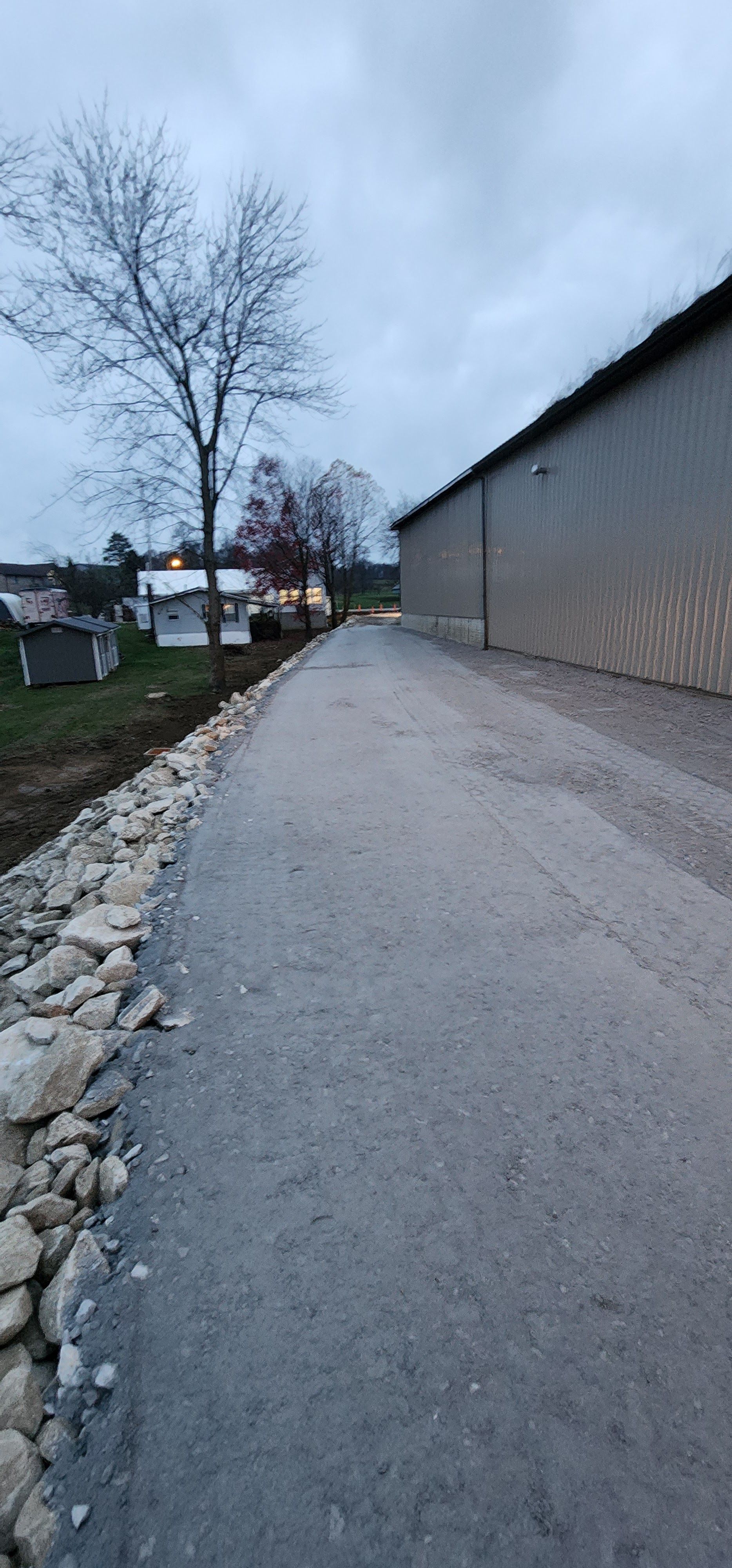 Gravel road next to a gray building and a stone border. Cloudy sky, bare tree, and grass in the distance.