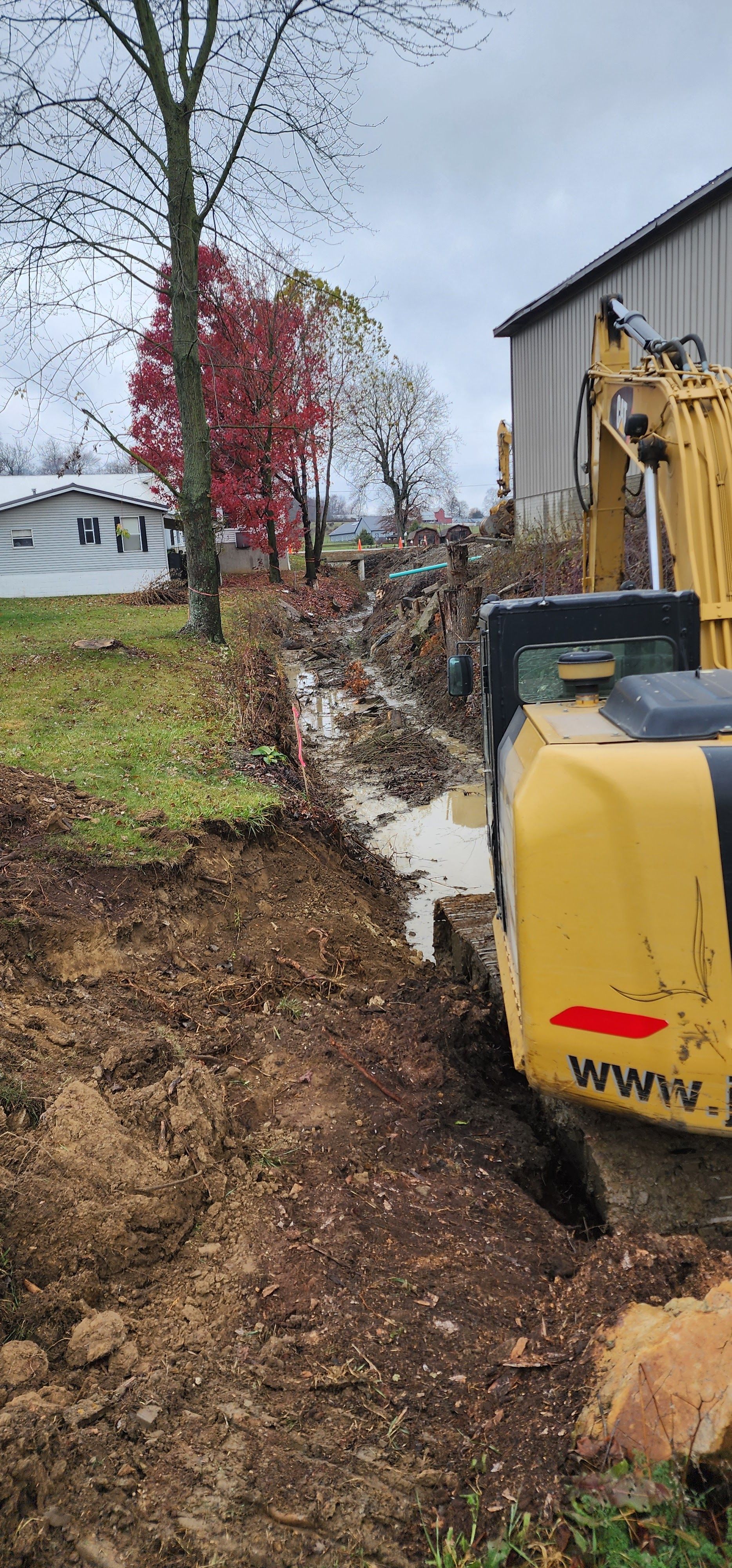 An excavator digging a trench next to a building, with a tree in the background. Cloudy day.