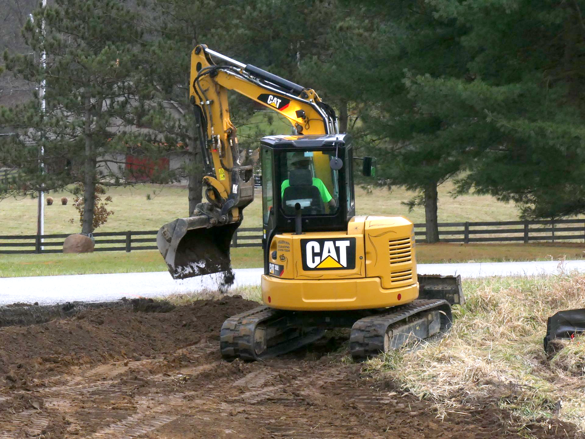 Yellow Caterpillar excavator digging in a muddy area near a fence and trees.