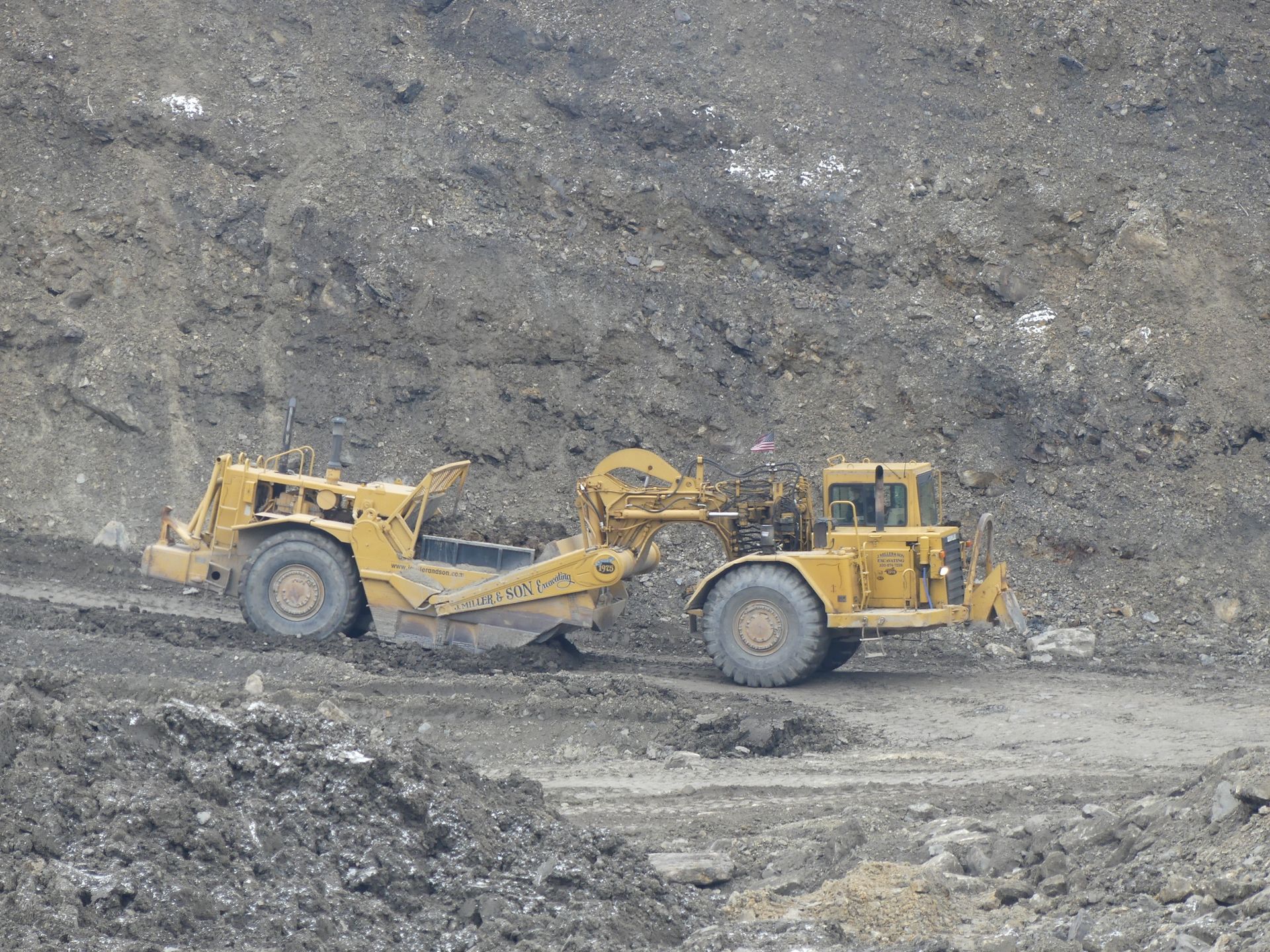 Yellow earthmover scraping earth from a hillside in an industrial setting.