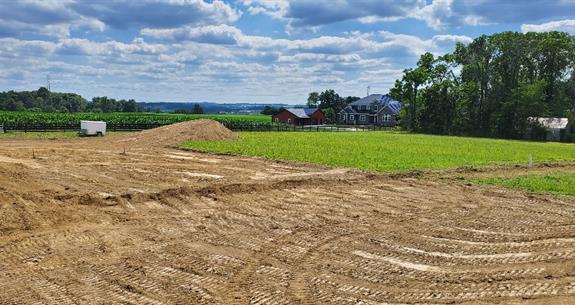 Brown earth in the foreground leads to green field, farmland, and cloudy sky. Buildings are visible in the distance.