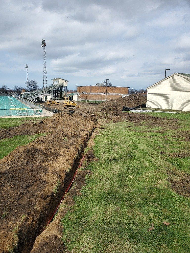 Trench dug in a grassy field, construction site in the background with buildings and heavy machinery. Overcast sky.