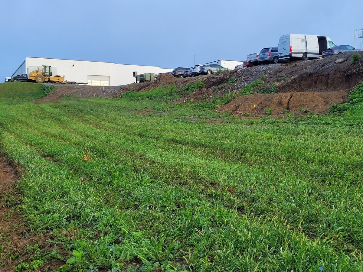 Green field slopes up to a pile of dirt and parked vehicles with a white building in the background.