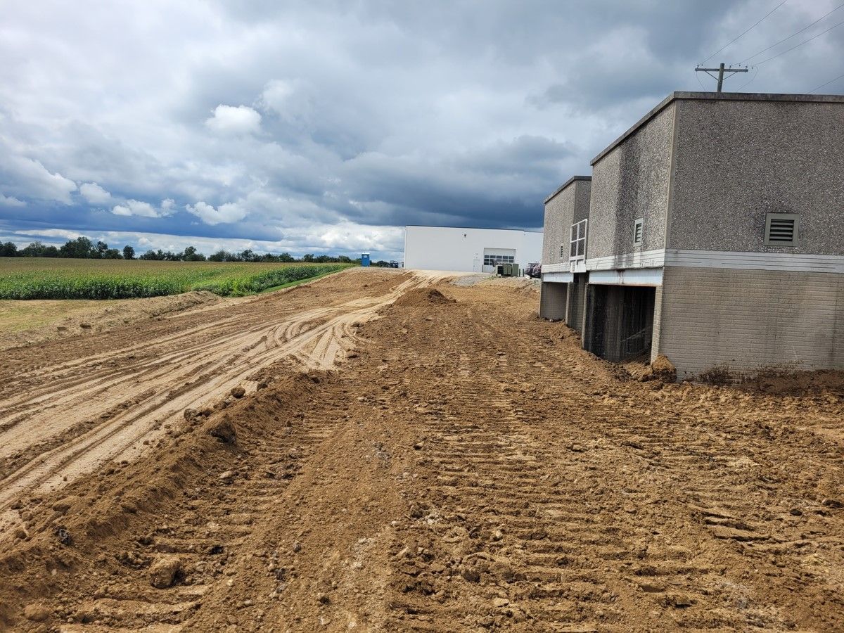 Construction site next to a field. Dirt mound with tire tracks, gray buildings, cloudy sky.