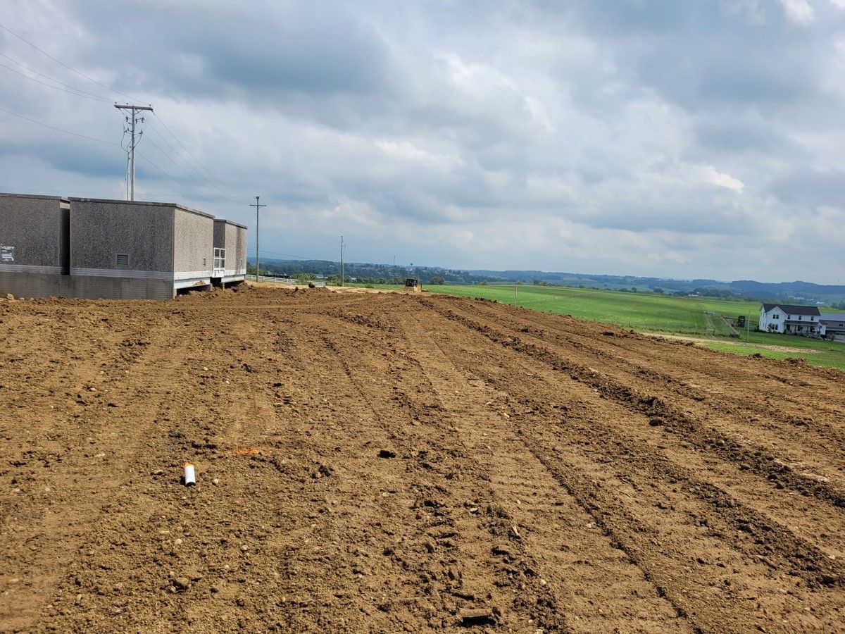 Brown earth with tire tracks in the foreground, incomplete brick buildings, and green fields under a cloudy sky.