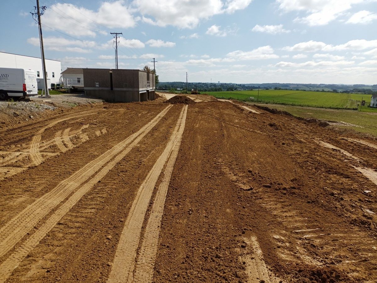 Muddy construction site with tire tracks, a few buildings, and fields in the distance under a blue sky.