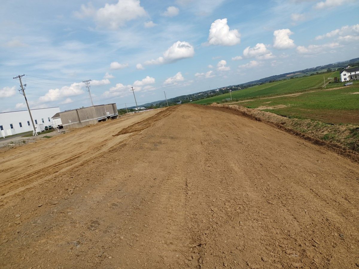 A large, freshly graded dirt path under a cloudy blue sky, next to a field and industrial buildings.