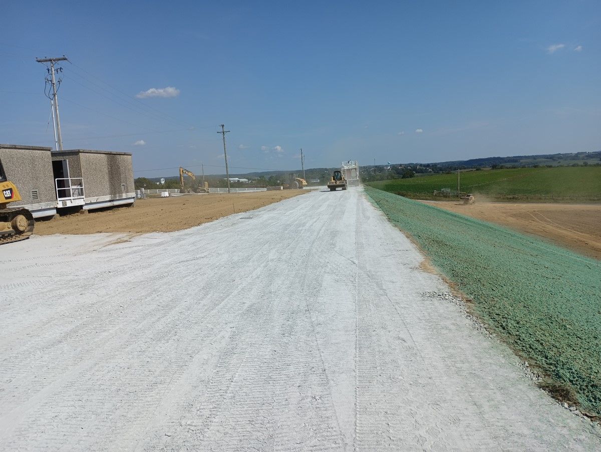 Road construction site with gravel surface, green erosion control, and heavy machinery under a blue sky.
