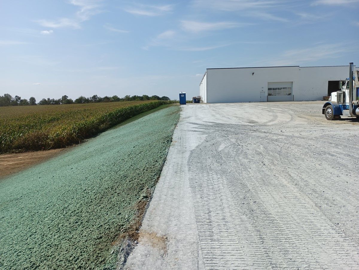 A gravel road transitions to a green-covered bank beside a field, building and truck in the distance under a blue sky.