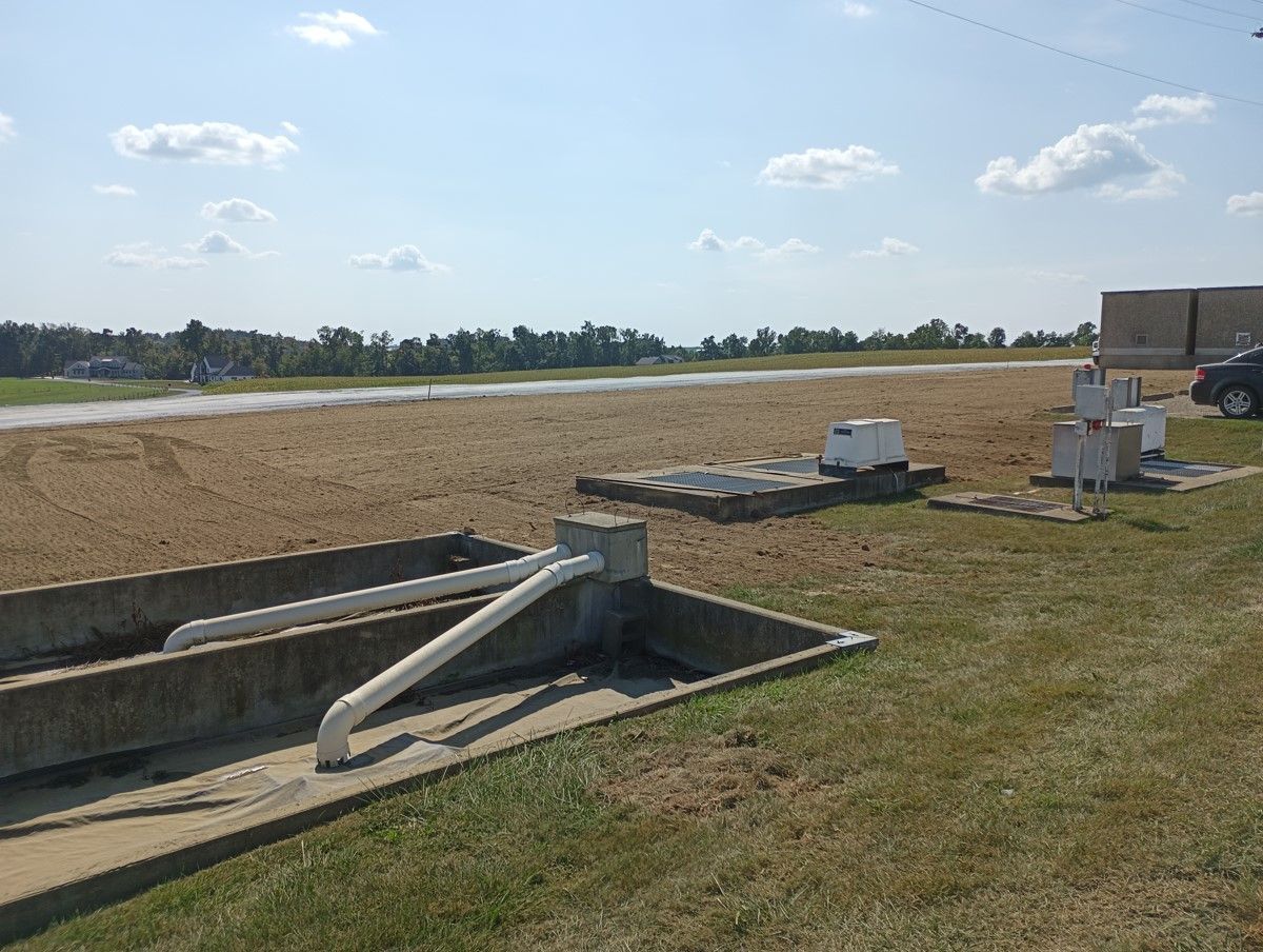Concrete tanks, pipes, and equipment on a grassy field; rural landscape in the background.