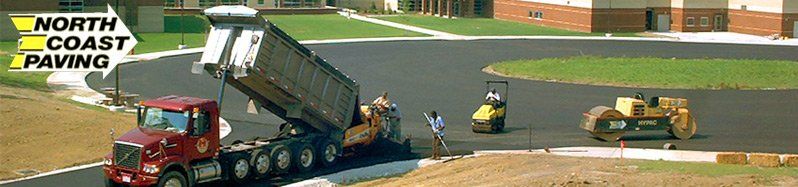 A dump truck unloads asphalt for paving, with a roller and compacting machine.