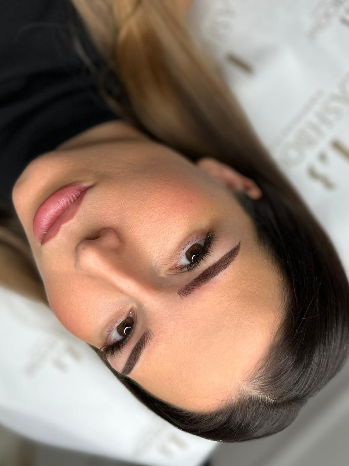 Woman with dark eyebrows and lip makeup, looking at camera. Lying on white surface.