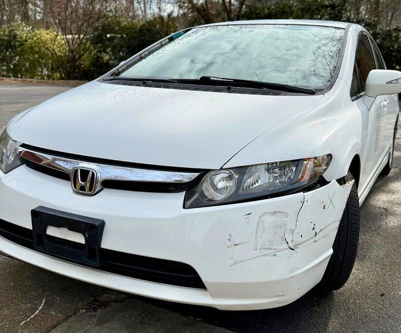 A white Honda Civic parked outdoors with noticeable scuffs and paint damage on the front bumper corner.