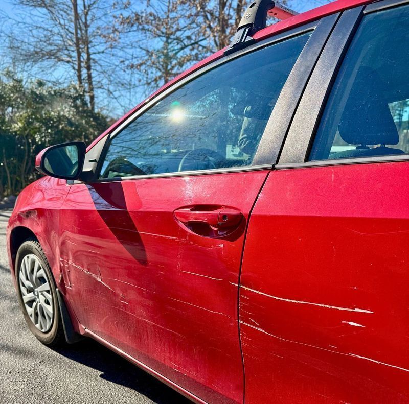 A red car parked outdoors with visible deep scratches and damage across the side door panels.