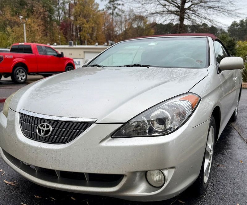 A silver Toyota Solara convertible parked on a wet pavement with a red pickup truck visible in the background.