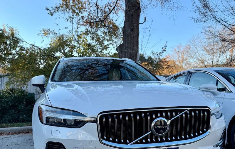 A white Volvo SUV parked outdoors, with a partial view of another white car next to it and trees in the background.